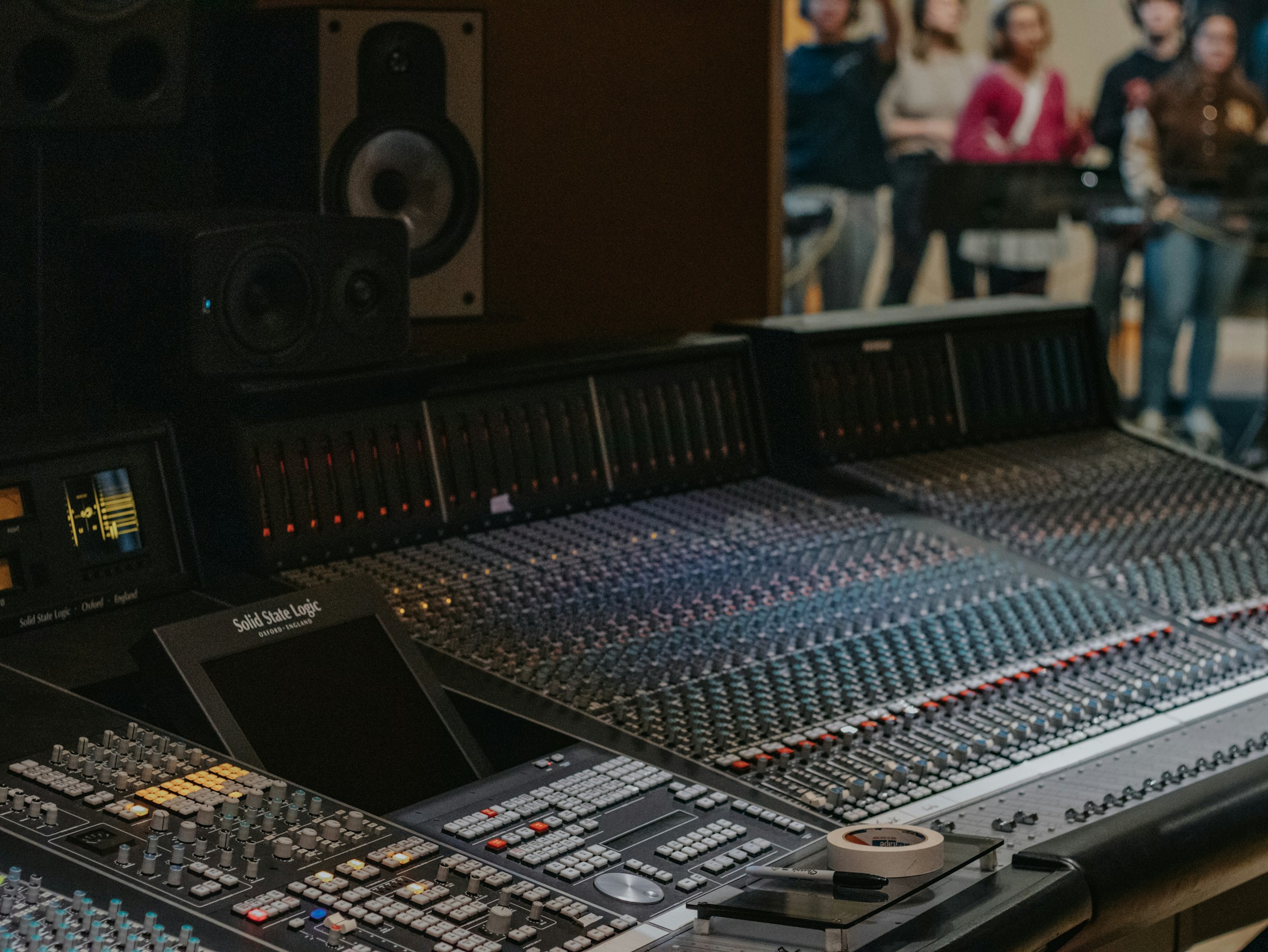 A studio mixing console with musicians visible beyond the control room
